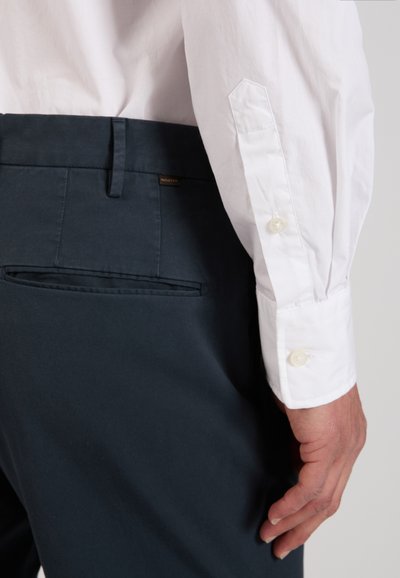 White long-sleeve shirt cuff with buttons and dark blue dress pants with back pocket and belt loop, shown from the side, on a standing person.