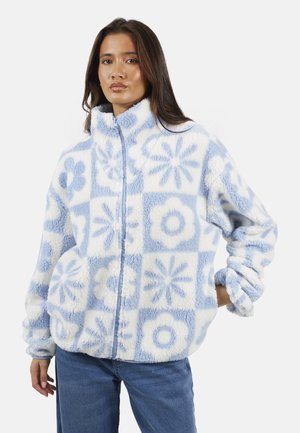 Donna che indossa una giacca di pile blu e bianca con motivi floreali, in piedi con una mano in tasca contro uno sfondo semplice.