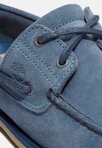 Niebieski zamszowy but z okrągłymi sznurowadłami, szarymi oczkami i kontrastującymi przeszyciami. Posiada wytłoczone logo z boku oraz miękką teksturę.