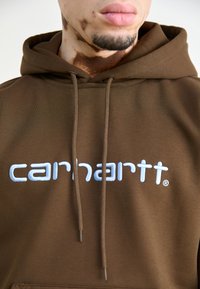 Brouna kapuce ar regulējamu auklu, ar baltu izšūtu "carhartt" logotipu, mīksta tekstūra un kangurū kabata.