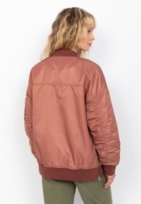 Chaqueta bomber de color rosa claro con cuello y puños acanalados, que presenta mangas texturizadas y un bolsillo lateral con cremallera. Acabado suave y similar al satén.