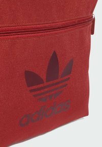 Borsa in tela rossa con un logo Adidas di un rosso mattone scuro e design a tre strisce. Include un comparto con zip sulla parte superiore.