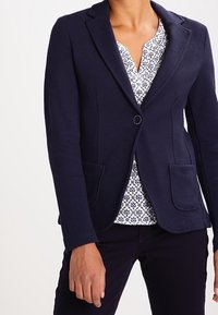 Námořnická texturovaná blazer s jedním knoflíkem, dvěma předními kapsami a klopovými límci, nošený přes vzorovanou blůzu.