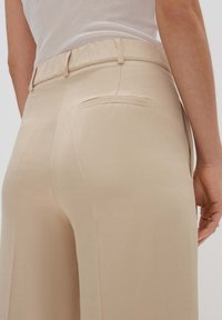 Pantalons beige à jambes larges avec une taille élastique, dotés d'une poche arrière unique et d'un tissu lisse et texturé.