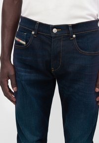 Homme portant un jean bleu foncé avec une chemise blanche, mains détendues sur les côtés, montrant les poches avant et la fermeture à boutons.