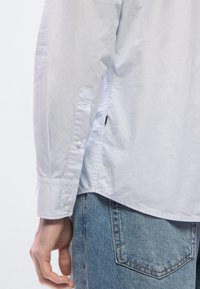 Vaaleansininen pitkähihainen nappipaita, jossa on hillitty tekstuuri ja sivuhalkiot, yhdistetty vaaleisiin farkkuihin.