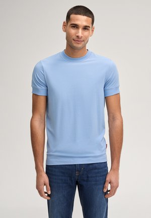 Man met kort donker haar, gekleed in een lichtblauw T-shirt met korte mouwen en donkerblauwe spijkerbroek, staand tegen een effen lichte achtergrond.