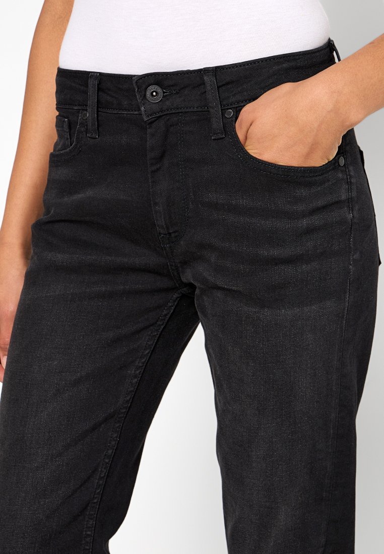 Jeans en denim noir avec une taille ajustée, design à cinq poches, texture subtile et fermeture à bouton. Une main repose dans la poche avant.