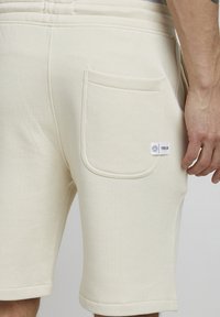 Helle beige Shorts aus weichem Stoff mit einer Gesäßtasche und einem Markenlabel. Sie verfügen über gestickte Details und einen lockeren Schnitt.