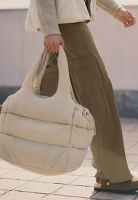 Sac fourre-tout en duvet beige avec une texture douce et des poignées rayées, associé à un pantalon vert olive avec des poches cargo.