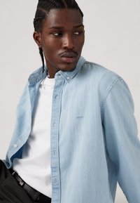 Cămașă din denim de culoare bleu deschis, cu guler tip buton, având detalii subtile de cusătură. Purtați peste un tricou alb, asortată cu pantaloni închiși la culoare.