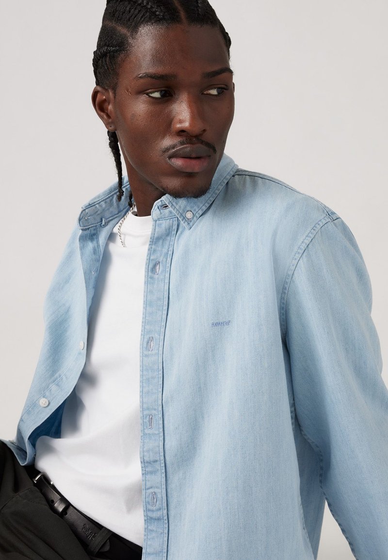Chemise en denim bleu clair avec un col boutonné, ornée de détails de couture subtils. Portée par-dessus un t-shirt blanc, associée à un pantalon sombre.