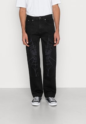 Vaqueros negros de pierna recta con patrones bordados de telarañas en la parte frontal de los muslos, combinados con zapatillas negras y blancas y una camiseta blanca.