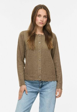 Femme aux longs cheveux bruns portant un cardigan marron boutonné et un jean bleu clair, debout devant un fond blanc uni.