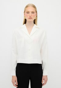 White cropped button-up shirt with a collar and single chest pocket, made of smooth fabric. Model wears black pants for contrast.