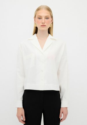 Weißes, kurzes Button-Down-Hemd mit Kragen und einer einzelnen Brusttasche, aus glattem Stoff gefertigt. Das Modell trägt schwarze Hosen für den Kontrast.