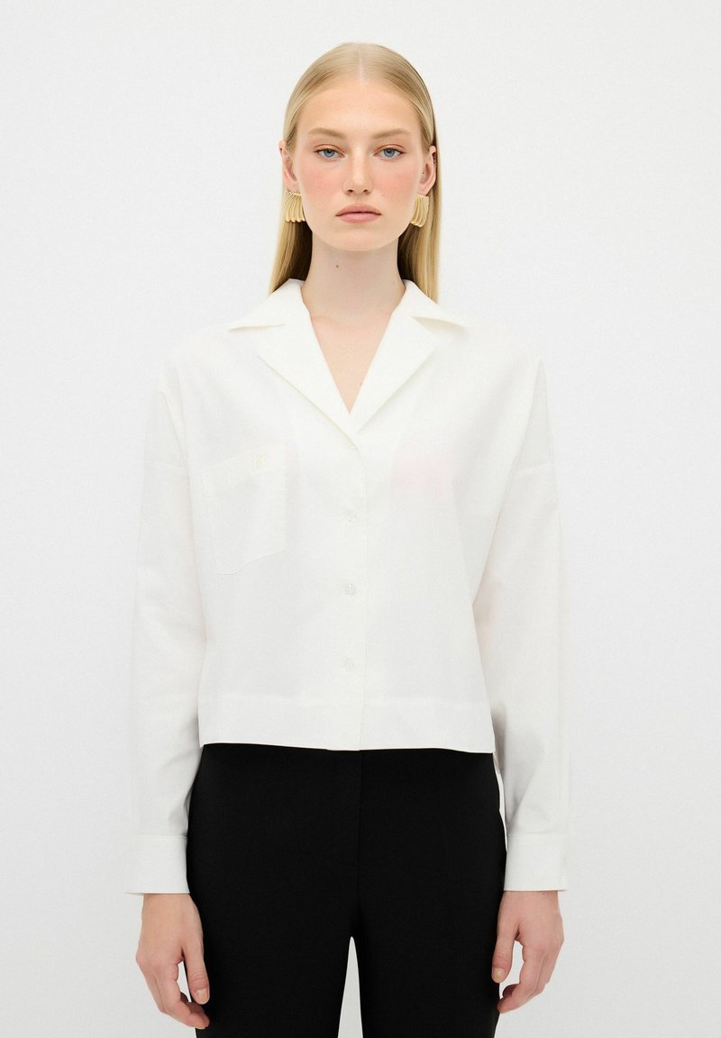 White cropped button-up shirt with a collar and single chest pocket, made of smooth fabric. Model wears black pants for contrast.