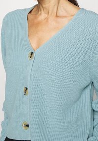 Hellblauer Strickcardigan mit tiefem V-Ausschnitt, großen runden Knöpfen und gerippter Textur. Details umfassen eine lockere Passform und betonte Ärmel.