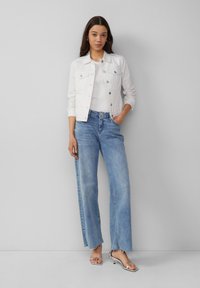 Witte denim jack met zilveren accenten over een witte ribbel top, gecombineerd met lichtblauwe high-waisted jeans met een rafelige zoom.