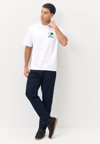 T-shirt bianca in cotone con logo verde "PADEL" e grafica di una racchetta. Abbinate a jeans scuri e scarpe in camoscio marrone.