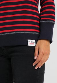Maglione a manica lunga a righe blu navy e rosso con una texture a costine, dettagli in cucitura ai polsini e un'etichetta visibile all'orlo. Sono indossati jeans neri.
