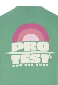 Žalias medvilninis marškinėliai, kuriuose yra didelė, daugiabriaunė grafika su vaivorykšte virš teksto "PRO TEST SUN SEA SURF" ryškiomis baltomis raidėmis.