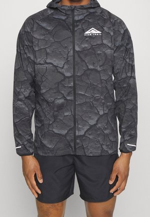 Mand iført sort Nike Trail hættetrøje med lynlås og revnet mønster samt sorte shorts, stående med hænderne afslappet langs siden.