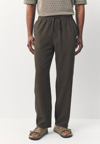 Brown, lightweight, woven pants with an elastic waistband and drawstring. Fitted straight style with side pockets. Worn with green slip-on sandals.