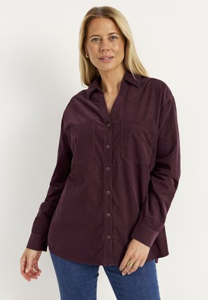Vrouw met blond haar, een donkerpaarse blouse met knopen en blauwe spijkerbroek, staand tegen een effen witte achtergrond.
