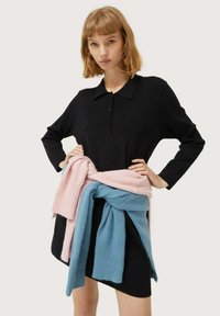 Vestido polo negro de manga larga con botones, llevado con suéteres de punto en tonos rosa y azul pastel anudados a la cintura. Textura suave, diseño casual.