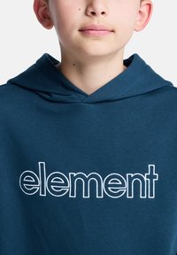 En ung gutt med lys hud har på seg en blå hettegenser med ordet "element" i hvit søm, og ser direkte inn i kameraet.