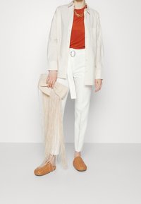 Witte slim-fit broek, oranje T-shirt, lichtbeige overhemd met knopen. Geweven tan instappers en een beige clutch met franjes. Gouden kettinghanger.