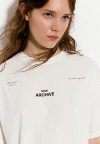 Valkoinen puuvillapaita, jossa on lyhyet hihat, pyöreä kaula-aukko ja mustat painetut tekstit: "UUSI ARKISTO", "Vaihtoehtoinen kulttuuri" ja "Kuurattu kappale."