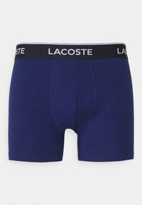 Jūras zila kokvilnas bokseršorti ar melnu jostasvieta, uz kuras uzraksts "LACOSTE" baltā krāsā. Gluda tekstūra, pieguļošs dizains un vidēja garuma griezums.