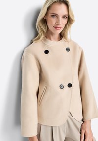 Giacca cropped in misto lana beige, con colletto rotondo, bottoni neri doppi e tasca laterale. Tessuto morbido, forma rilassata.