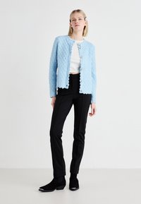 Light blue textured cardigan with scalloped edges and round buttons, paired with black slim trousers and black ankle boots.