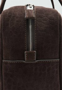 Mochila de ante marrón con un acabado texturizado, que presenta un zipper plateado, costuras marrones y un bolsillo plano en la parte frontal.