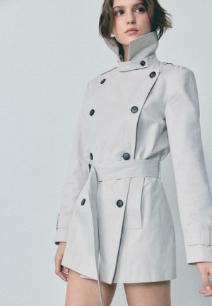Femme portant un trench-coat gris clair à double boutonnage avec un col montant et une ceinture, debout devant un fond uni.