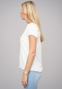 T-shirt bianco a maniche corte, con dettagli in pizzo testurizzato sulle spalle, vestibilità morbida e tessuto soffice. Indossato con jeans blu chiaro.
