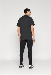 Μαύρο πόλο μπλουζάκι με κοντά μανίκια, συνδυασμένο με μαύρο παντελόνι στενής γραμμής και αθλητικά παπούτσια Adidas με τρεις λευκές ρίγες.