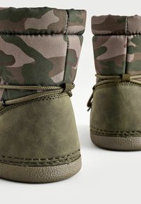 Des bottes de cheville vertes en camouflage avec une tige en tissu texturé et une partie inférieure en cuir lisse, dotées de accents cousus et d'une semelle en caoutchouc ronde.