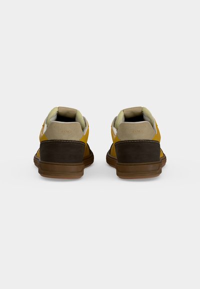 Paire de baskets jaunes et marron vues de dos avec des cols rembourrés beige et des semelles en caoutchouc marron sur un fond gris uni.