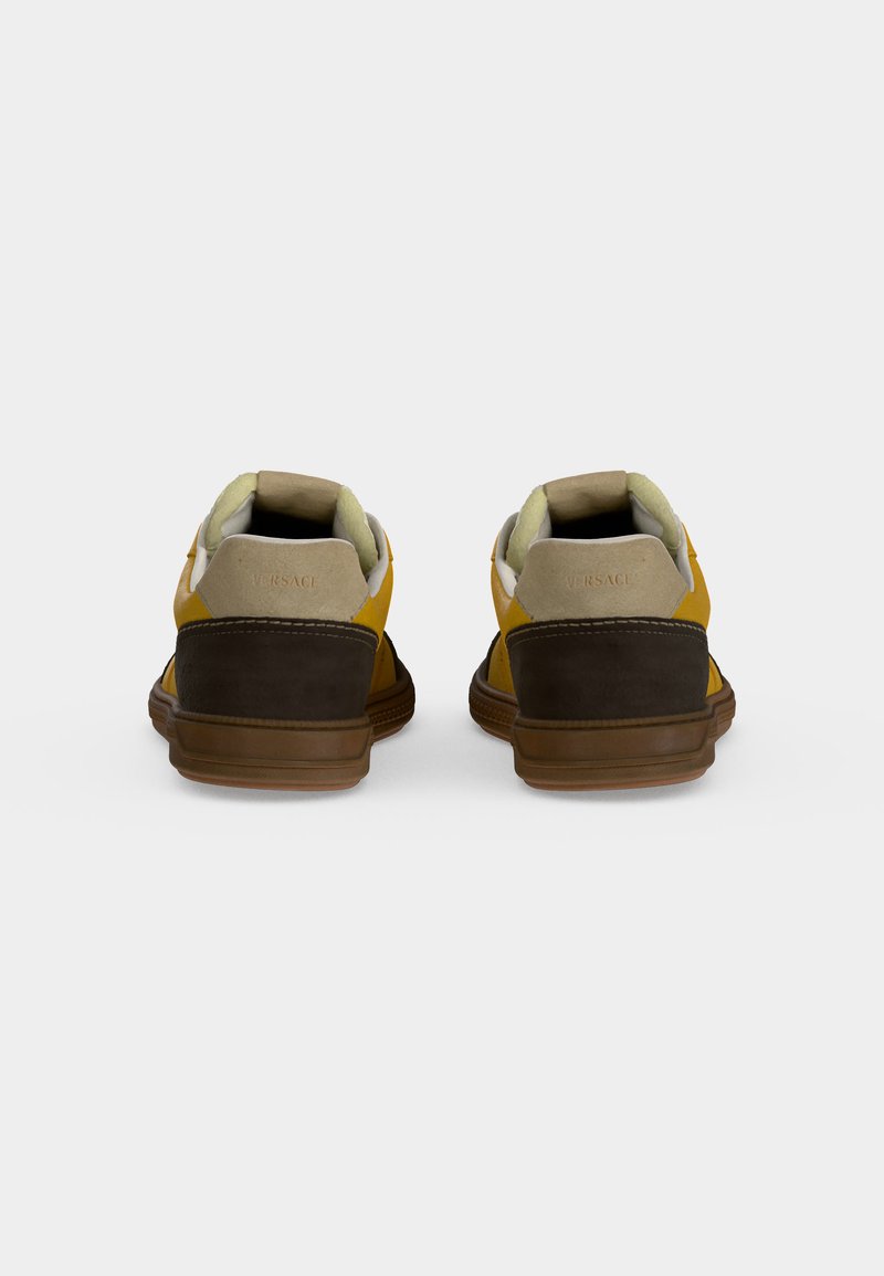 Coppia di sneakers gialle e marroni viste da dietro con colli imbottiti beige e suole in gomma marrone su uno sfondo grigio semplice.