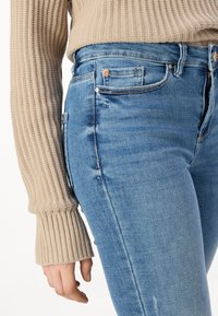 Vaaleansiniset farkut, joissa on slim fit -malli, viisi taskua, oranssia ommeltyötä ja ribbikuvioinen vaaleanbeige villapaita pitkillä hihoilla.