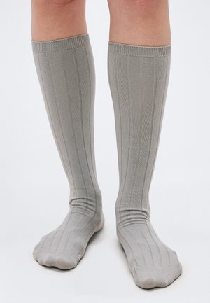 Πόδια που φορούν ανοιχτό γκρι ριγωτές κάλτσες μέχρι το γόνατο, στεκόμενα σε λευκή επιφάνεια με τα πόδια ελαφρώς ανοιχτά.