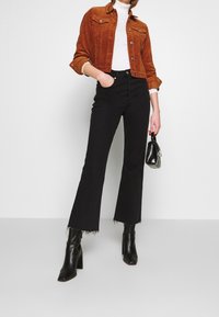 Vrouw in een roestkleurige corduroy jas, witte turtleneck, zwarte cropped flare jeans en zwarte hoge hakken, met een kleine zwarte handtas.