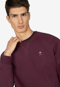Sudadera burdeos con un cuello redondo azul marino, que presenta un pequeño diseño bordado en el pecho y costuras texturizadas visibles.