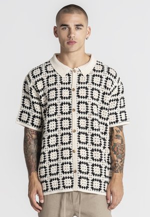 Junger Mann mit kurzen Haaren und Tätowierungen, der ein schwarz-weiß gemustertes kurzärmliges Hemd mit Knöpfen und beige Hosen trägt, blickt nach vorne.