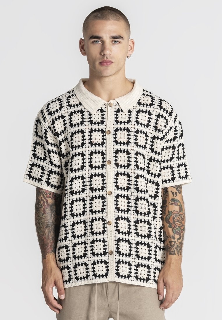 Mladić s kratkom kosom i tetovažama, odjeven u crno-bijelu košulju s kratkim rukavima s uzorkom te bež hlače, okrenut prema naprijed.