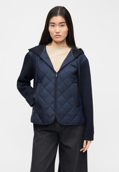 Femme aux longs cheveux noirs portant une veste matelassée bleu marine à fermeture éclair avec capuche et un pantalon noir à jambes larges, debout devant un fond uni.